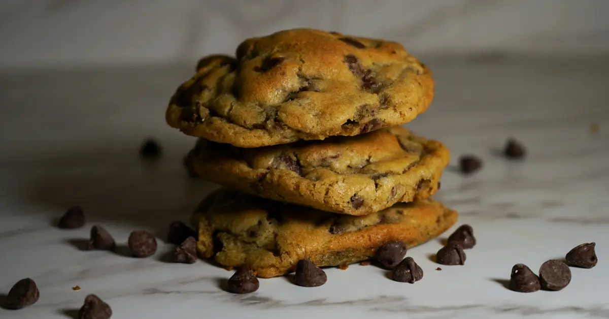 A stack of freshly baked Nestle chocolate chip cookies on a rustic wooden table, with melty chocolate chips and a glass of milk in the background.